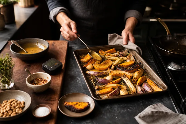 A cook bastes roasted vegetables beside measured ingredients and a timer.